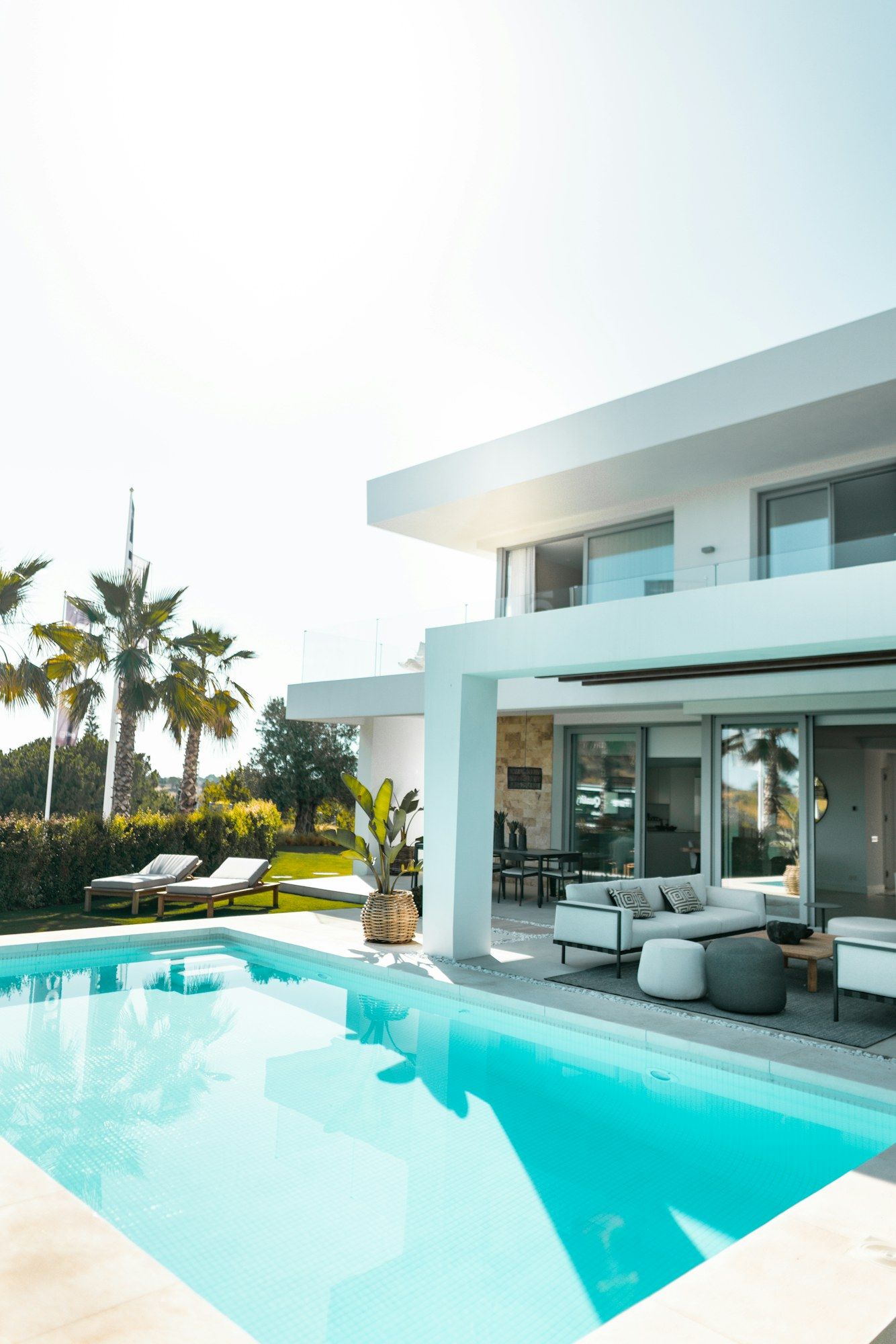 A modern white two-story home with a turquoise swimming pool, a shaded patio with outdoor seating, and palm trees under a bright sky